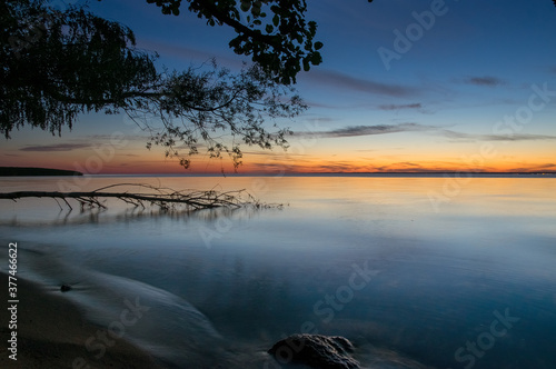 Krajobraz zachód słońca nad wodą z pięknie oświetlonym niebem i starym drzewem powalonym do wody © Monika