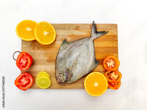 top view of Black Pomfret Fish dish cooking with various ingredients. Fresh raw fish decorated with lemon slices and tomato slice and curry leaves on a wooden pad,white background. photo