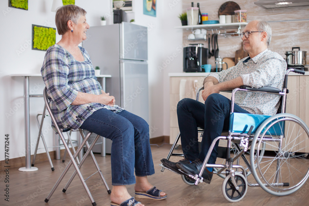 Old woman and her disabled husband in wheelchair chatting in kitchen.Elderly person having a conversation with husband in kitchen. Living with disabled person with walking disabilities Old woman and her disabled husband in wheelchair chatting in kitchen.Elderly person having a conversation with husband in kitchen. Living with disabled person with walking disabilities