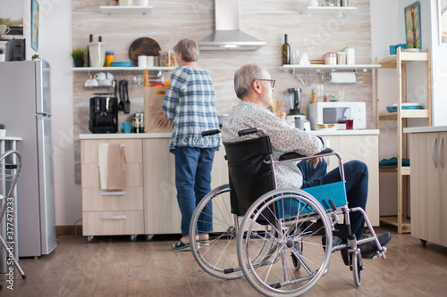 Pensive disabled elderly person in wheelchair looking on the window from the kitchen. Disabled man sitting in wheelchair in kitchen looking through window while wife is preparing breakfast. Invalid