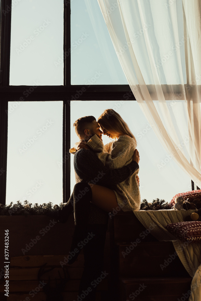 Silhouette of couple hugging in front of a big panoramic window with bright blue sky on the background.