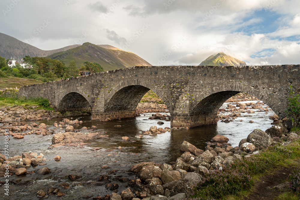 Old stone arch bridge over a mountain river at Sligachan on the Isle of Skye in the Highlands of Scotland, the Cuillin mountains rising behind lit by sunset