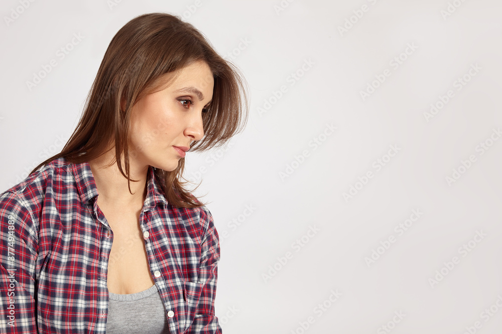 Young beautiful woman with dark hair and eyes, wearing casual checkered shirt and gray t-shirt, looks down with serious face expression. Dreaming mood, natural make-up. Light background, copy space. Young beautiful woman with dark hair and eyes, wearing casual checkered shirt and gray t-shirt, looks down with serious face expression. Dreaming mood, natural make-up. Light background, copy space.