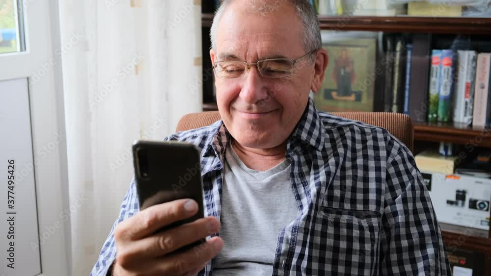 an elderly man sits in a chair with a smartphone in his hand and laughs an elderly man sits in a chair with a smartphone in his hand and laughs