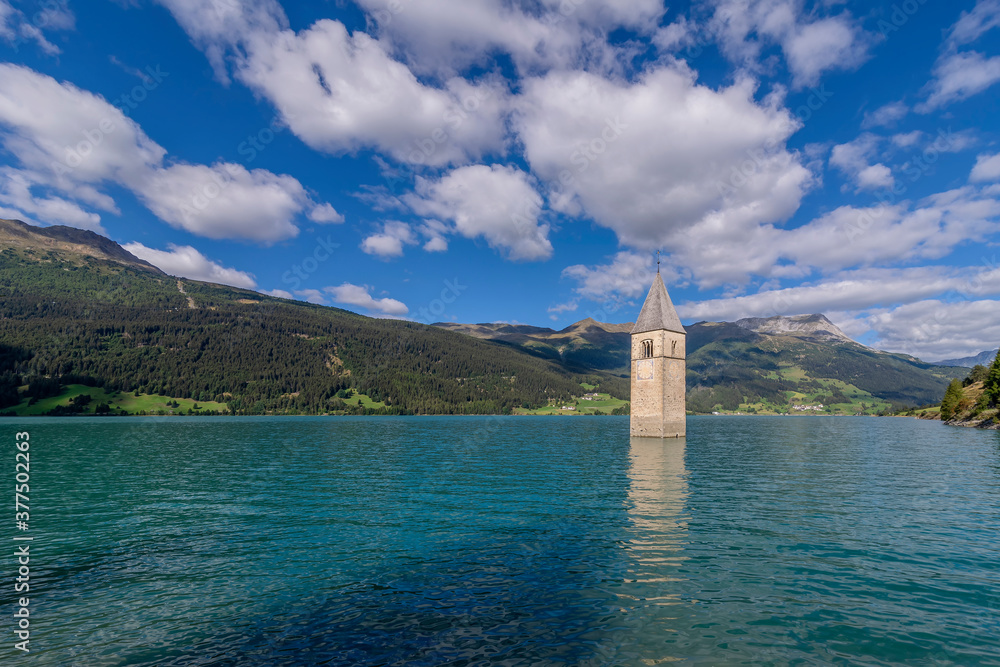 The ancient submerged bell tower of Lake Resia in Val Venosta, South Tyrol, Italy, emerges from the water against a beautiful blue sky with white clouds