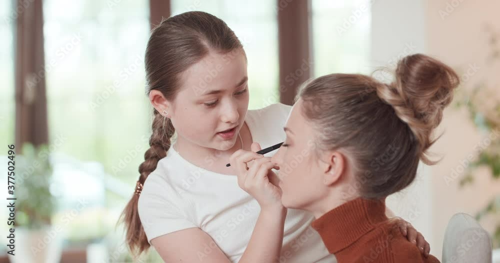 Teen girl is learning to apply make up to her mother - she applies the eyshadows on the eyelids of mother, and they are talking. Window, white wall and green plants at background