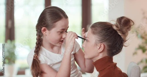 Teen girl is learning to apply make up to her mother - she applies the eyshadows on the eyelids of mother, and they are talking, and smiling. Window, white wall and green plants at background photo