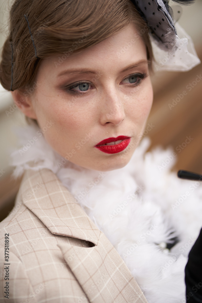 Beautiful brunette woman in a hat, black retro dress with red lips, beige coat. Sitting on a bench looking away