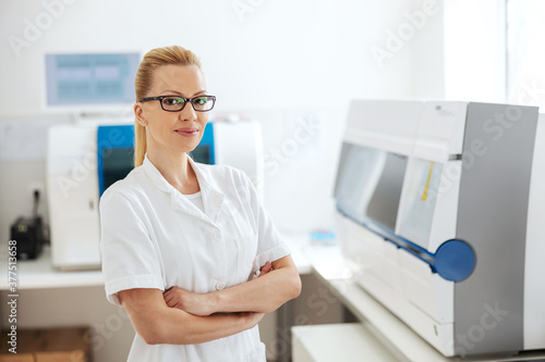 Beautiful blond female lab assistant with eyeglasses standing in lab with arms crossed and looking at camera.