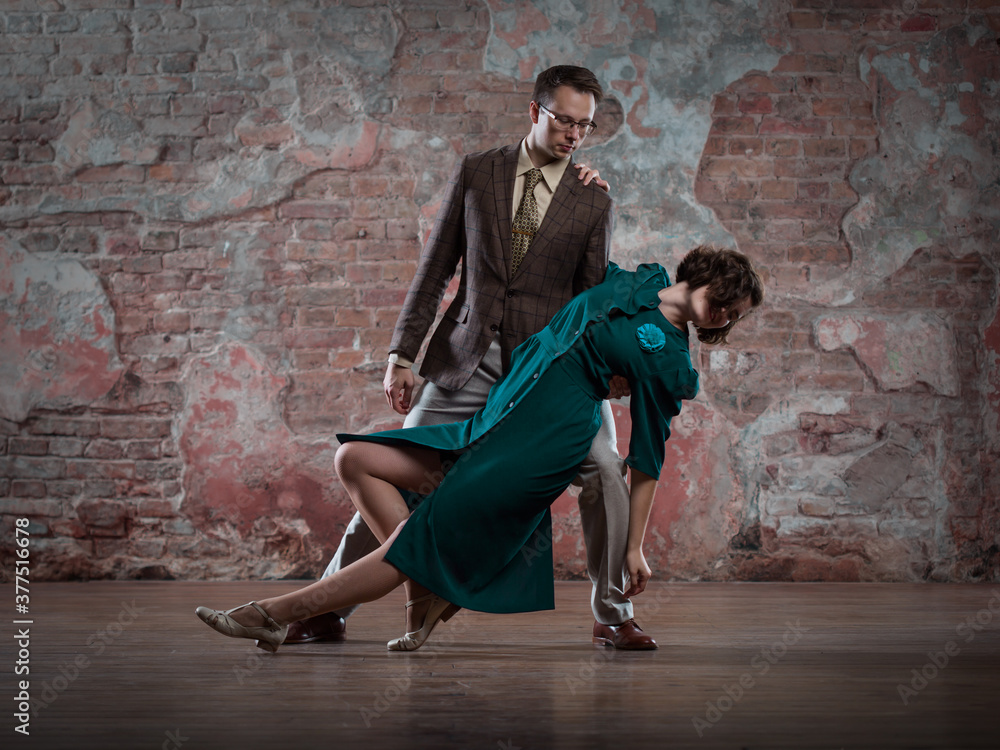a young couple - a man in a suit and a woman in a dark-turquoise dress - dancing against an old brick wall
