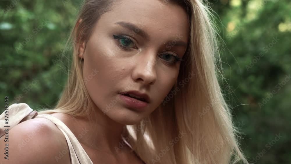 Young woman with long loose flowing fair hair poses running hand on head, and looking at bare shoulder against blurred dense green forest slow motion close view Young woman with long loose flowing fair hair poses running hand on head, and looking at bare shoulder against blurred dense green forest slow motion close view