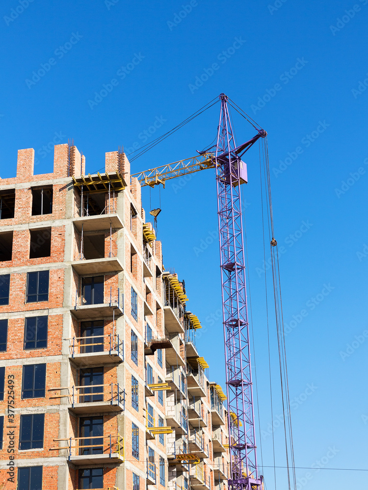 Crane near the brick high-rise building under construction against the blue sky, modern construction Crane near the brick high-rise building under construction against the blue sky, modern construction