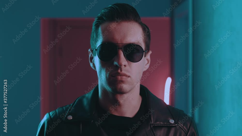 Portrait of brutal man inside at night over neon lights of nightclub. Serious male biker put on sunglasses indoors on blue and pink neon light background Portrait of brutal man inside at night over neon lights of nightclub. Serious male biker put on sunglasses indoors on blue and pink neon light background