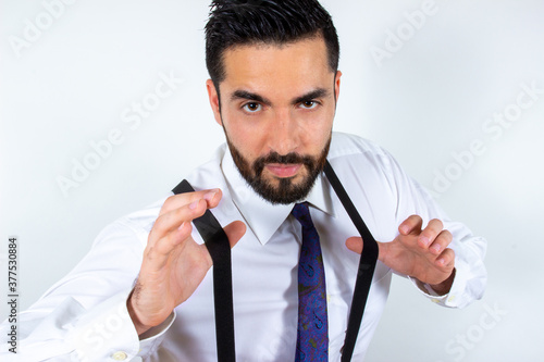 Handsome young businessman pulling his suspenders, looking at camera, superiority expression. Close-up. Portrait of attractive stylish bearded man in formal wear. White background.