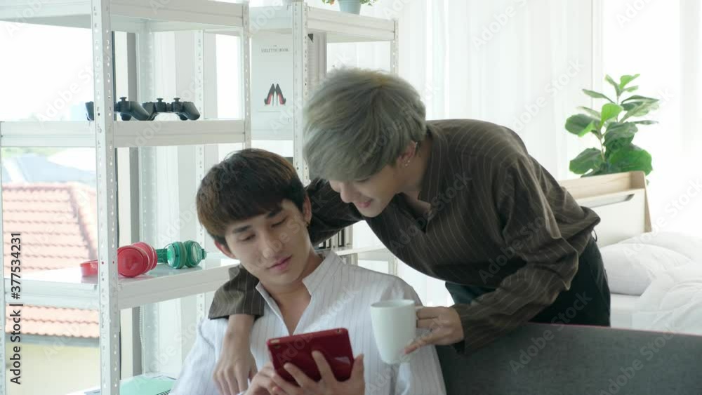 Relax and romantic LGBT couple in daily-life concept. Handheld medium shot of Asian men in living room. One using laptop tablet another holding a cup of coffee. They cuddle, smile, make eye contact Relax and romantic LGBT couple in daily-life concept. Handheld medium shot of Asian men in living room. One using laptop tablet another holding a cup of coffee. They cuddle, smile, make eye contact
