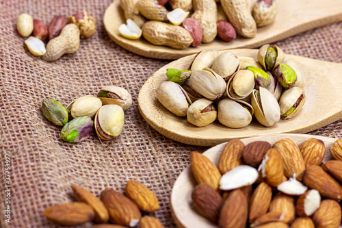 Different nuts: almonds, pistachios, peanuts, hazelnuts in light wooden spoons on beige textured textile background.