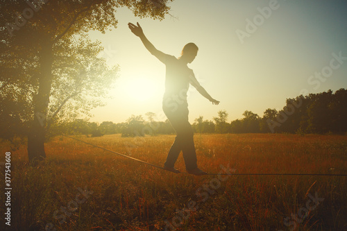  Young man walking on slackline in the meadow at sunset.