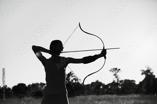 Fotografering Young Caucasian female archer shooting with a bow in a field at sunset
