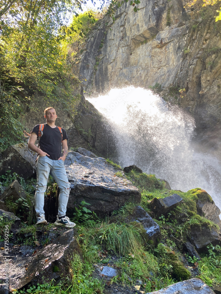 Portrait of young man looking away on background of waterfall. Adult male tourist enjoying beautiful view in mountainous area Portrait of young man looking away on background of waterfall. Adult male tourist enjoying beautiful view in mountainous area