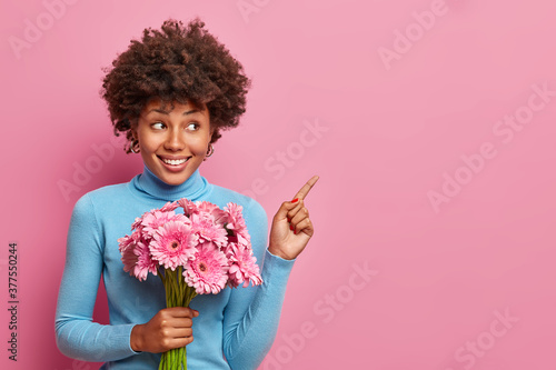 Smiling Afro American lady has happy mood and points at upper right corner, holds bouquet of gerbera flowers during her birthday. Beautiful woman shows direction to flowers shop, pink background
