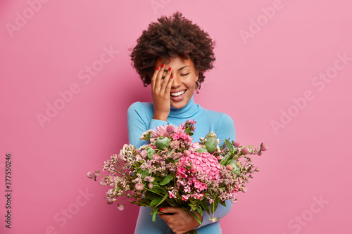 Joyful optimistic woman with Afro hair makes face palm and giggles positively, holds nice bouquet received from beloved person. Happy lady celebrates spring holiday, enjoys beautiful flowers