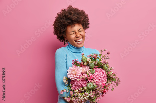 Portrait of happy young African American woman laughs joyfully, closes eyes and poses with beautiful bouquet of flowers. Positive female model has fun during holiday, expresses positive emotions