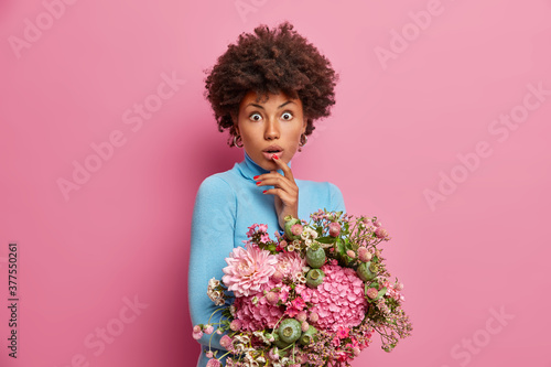 Isolated shot of astonished curly haired woman with natural makeup holds nice flowers, shocked to get unexpected delivery on special occasion. Impressed birthday woman carries beautiful bouquet
