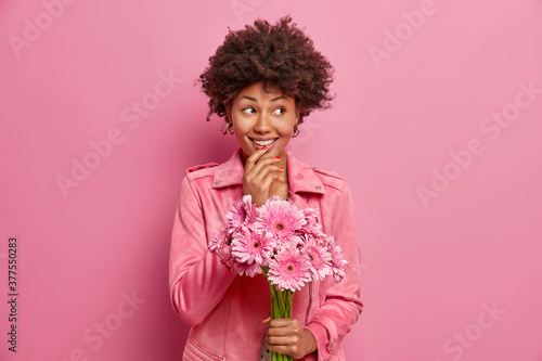 Positive African American woman with toothy smile holds bouquet of beautiful flowers, looks aside and wears fashionable jacket. Happy birthday girl gets rosy gerbera daisy from beloved person