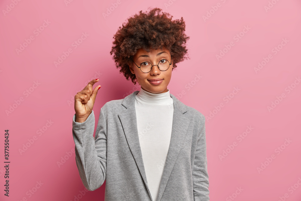 Displeased curly haired woman entrepreneur in formal clothes shows something very tiny or small, demonstrates size of salary and stands against pink background. Measure and body language concept Displeased curly haired woman entrepreneur in formal clothes shows something very tiny or small, demonstrates size of salary and stands against pink background. Measure and body language concept