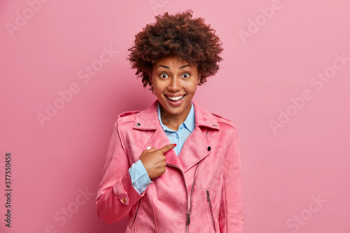 Happy African American woman points at herself and asks who me, wears fashionable clothes. Joyful ethnic lady being picked by someone, isolated over pink background. Girlfriend asked to participate