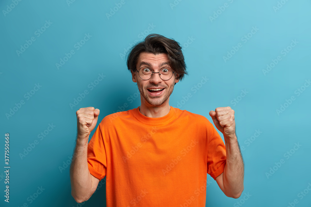 Fototapeta premium Hopeful man supports favorite football team, raises clenched fists and looks with excitement at camera. Thrilled amused guy in orange t shirt fascinated to win first prize isolated on blue studio wall