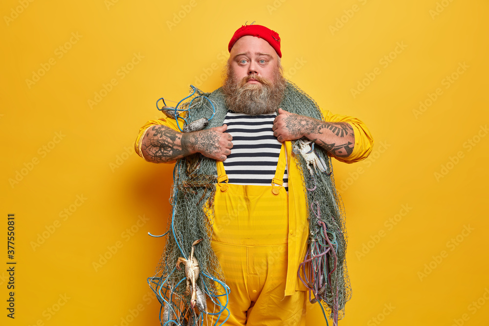 Serious plump bearded experienced man sailor poses with fishing gears, wears striped sailor shirt and overalls. Fisherman has sea adventure, poses indoor against yellow background. Hobby concept