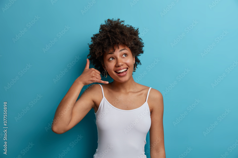 Happy dreamy dark skinned woman shows call me gesture, smiles broadly and dressed casually. Cheerful African American millennial girl asks for telephone number, poses against blue studio wall Happy dreamy dark skinned woman shows call me gesture, smiles broadly and dressed casually. Cheerful African American millennial girl asks for telephone number, poses against blue studio wall