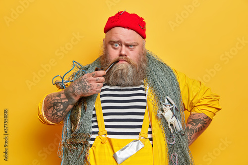 Close up shot of serious bearded sailor smokes pipe and looks unhappily at camera, poses with fishing net against yellow studio wall. Angry tattooed fisherman in overalls spends free time at sea