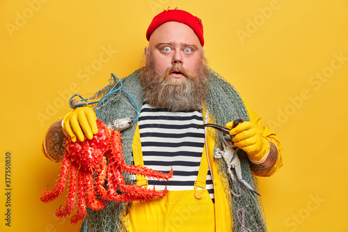 Stupefied bearded man shocked to catch big octopus during fishing time, smokes pipe and stares at camera. Fisherman with fish net enjoys leisure activity or hobby, stands indoor over yellow wall photo