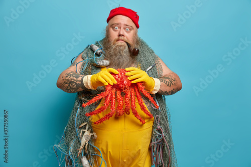 Isolated shot of bearded plump man smokes pipe and holds red octopus, poses with fishing net after successful angling on seashore. Thick shocked male sailor caught sea creature during marine voyage