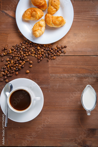 A mug of coffee and a jug of cream and croissants on the table among the scattered coffee beans.