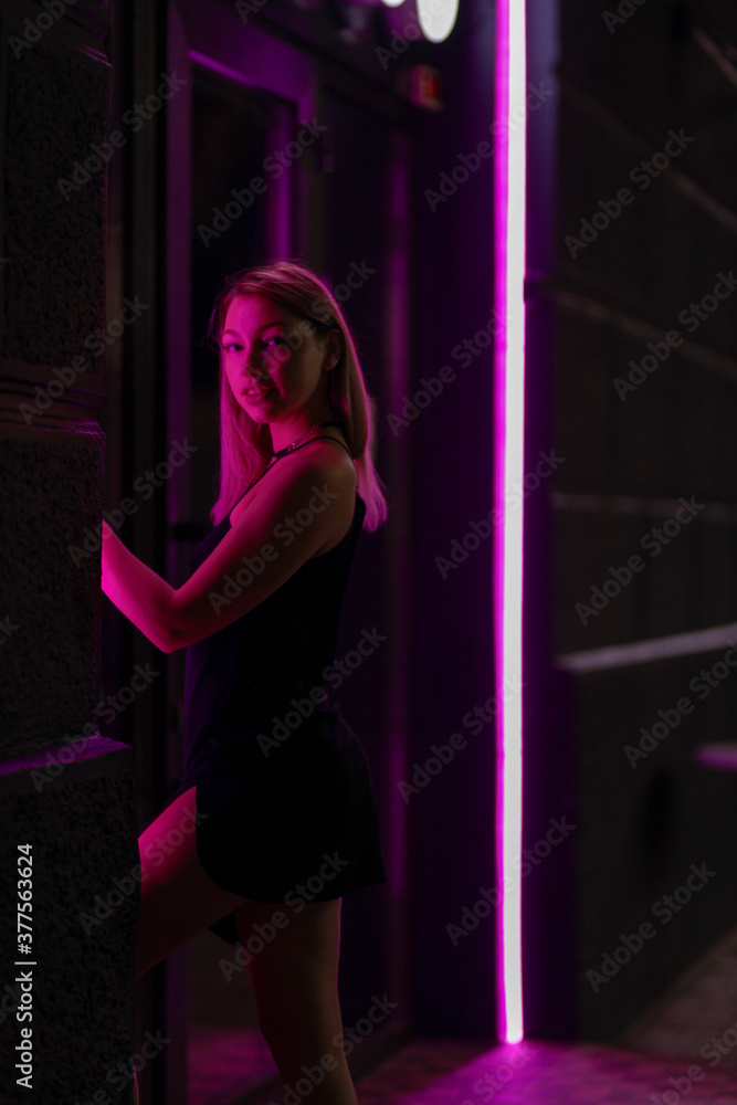 Club style photo of girl in a black dress. Set is lit with violet light. Picture has dark night tone. She stands at the entrance to the building Club style photo of girl in a black dress. Set is lit with violet light. Picture has dark night tone. She stands at the entrance to the building
