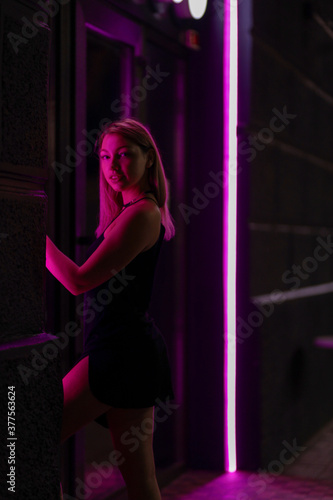 Club style photo of girl in a black dress. Set is lit with violet light. Picture has dark night tone. She stands at the entrance to the building