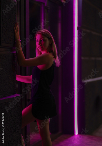 Club style photo of girl in a black dress. Set is lit with violet light. Picture has dark night tone. She stands at the entrance to the building