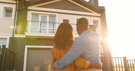 Rear on African American young just-married couple standing at yard of house, hugging and observing second floor. Back view on man and woman in hugs moving in new home.