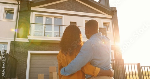 Murais de parede Rear on African American young just-married couple standing at yard of house, hugging and observing second floor