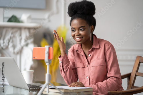 African American millennial woman with afro hairstyle remote studying, working online on laptop, chatting with friends via video call on smartphone on tripod. Blogger influencer recording video blog. photo