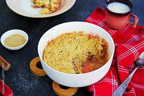 Dessert, crispy crumble with rhubarb in a white glass mold on a dark concrete background. Served with custard.