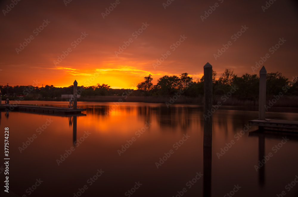 Sunset over a river, creating silhouette of docks and pilings, as well as trees.