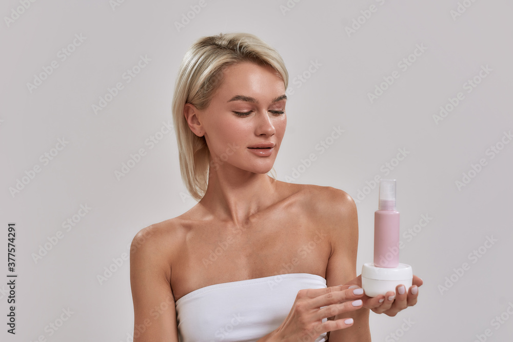 Portrait of young woman with perfect glowing skin holding jar and bottle of different cosmetic skincare products and looking at it while posing isolated over grey background Portrait of young woman with perfect glowing skin holding jar and bottle of different cosmetic skincare products and looking at it while posing isolated over grey background