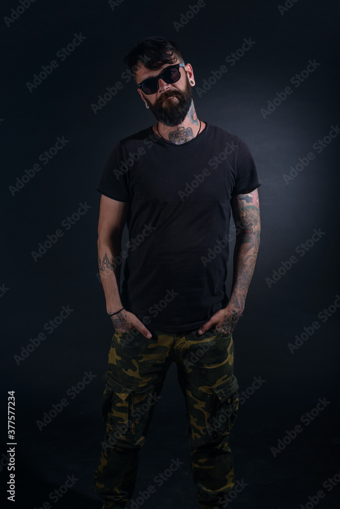 Bearded male model with tattoos and hands in his pocket, dressed in a black t-shirt and sunglasses poses over black background. Bearded male model with tattoos and hands in his pocket, dressed in a black t-shirt and sunglasses poses over black background.