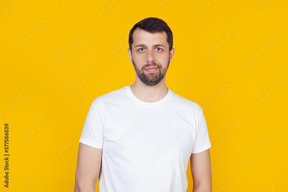 Young man with a beard in a white t-shirt happy face smiling looking at the camera. Positive man standing on isolated yellow background Young man with a beard in a white t-shirt happy face smiling looking at the camera. Positive man standing on isolated yellow background