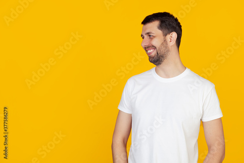 Young man with a beard in a white t-shirt looking to the side with a smile on his face, natural expression. Laughs confidently, stands on isolated yellow background
