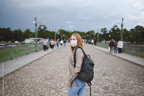 Young blonde woman walking in city street wearing face protective mask for Covid 19 prevention. Caucasian young student or traveler tourist among the crowd with backpack. Corona virus concept © Алина Троева